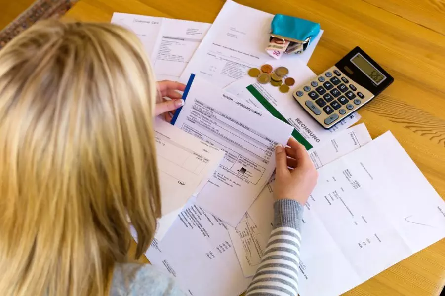 blonde woman looking at bills, papers and a calculator on a wooden table
